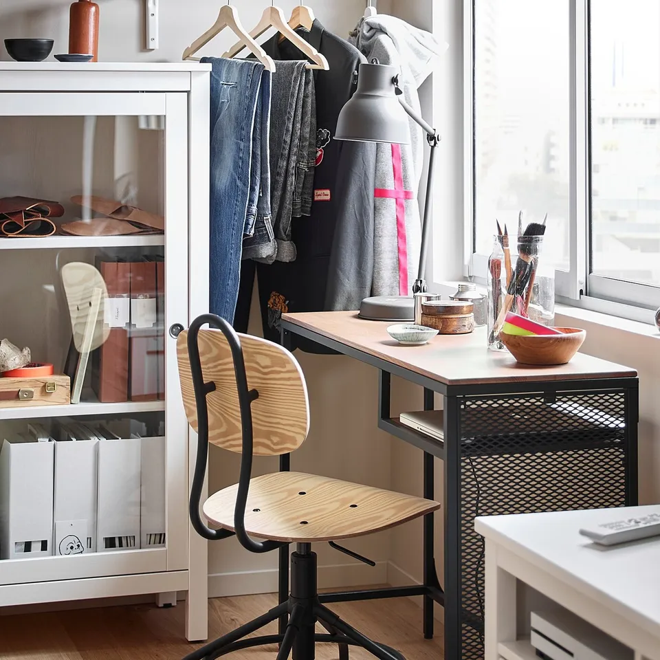 Modern home office: wooden chair, black metal base, white desk, clothes rack, bookshelf, art supplies, wood floor.