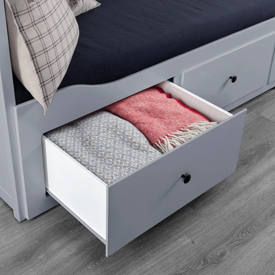 White daybed drawer open, showing red and grey blankets inside, with black handles and a grey plaid pillow on the bed.