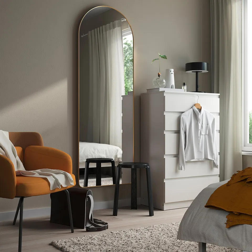Minimalist bedroom with gold mirror, dresser, orange chair, bed with grey & mustard linens.