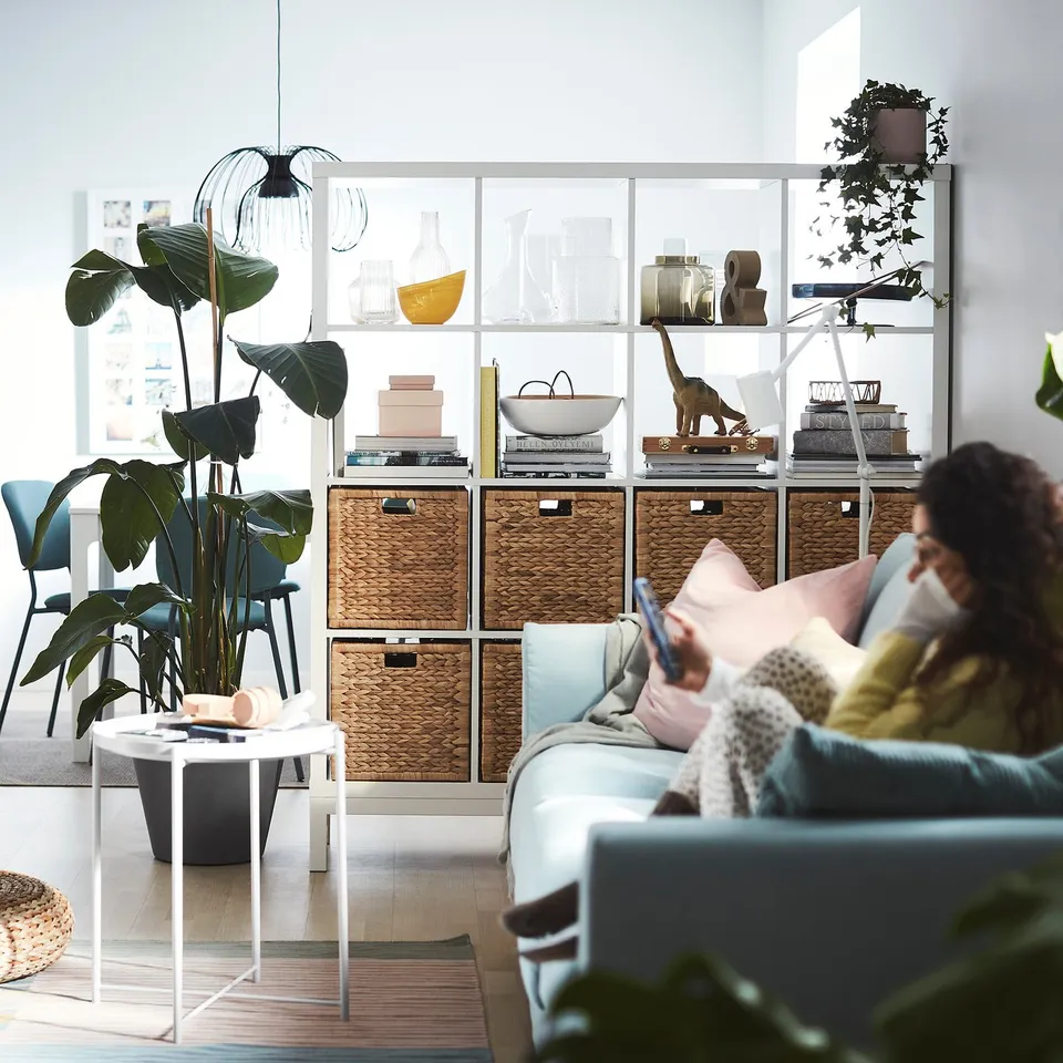 Living room with blue sofa, coffee table, KALLAX shelves, books, decor, person on smartphone.