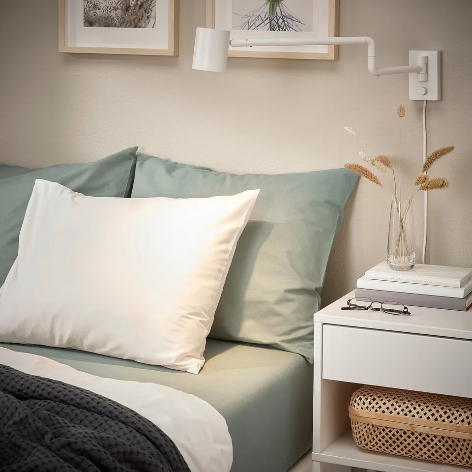 Minimalist bedroom: gray-green fitted sheet on bed, white pillows, small bedside table with books, glasses, vase, and woven box.