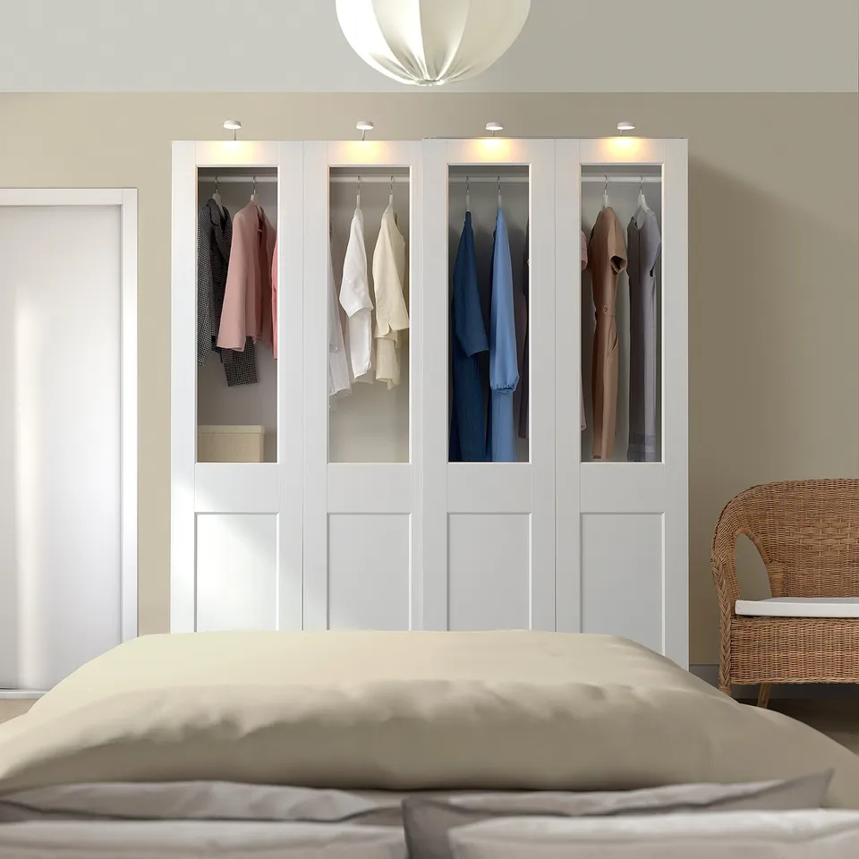 Modern bedroom with large white PAX wardrobe, colourful clothes, integrated lighting, cosy beige bed, and wicker chair.