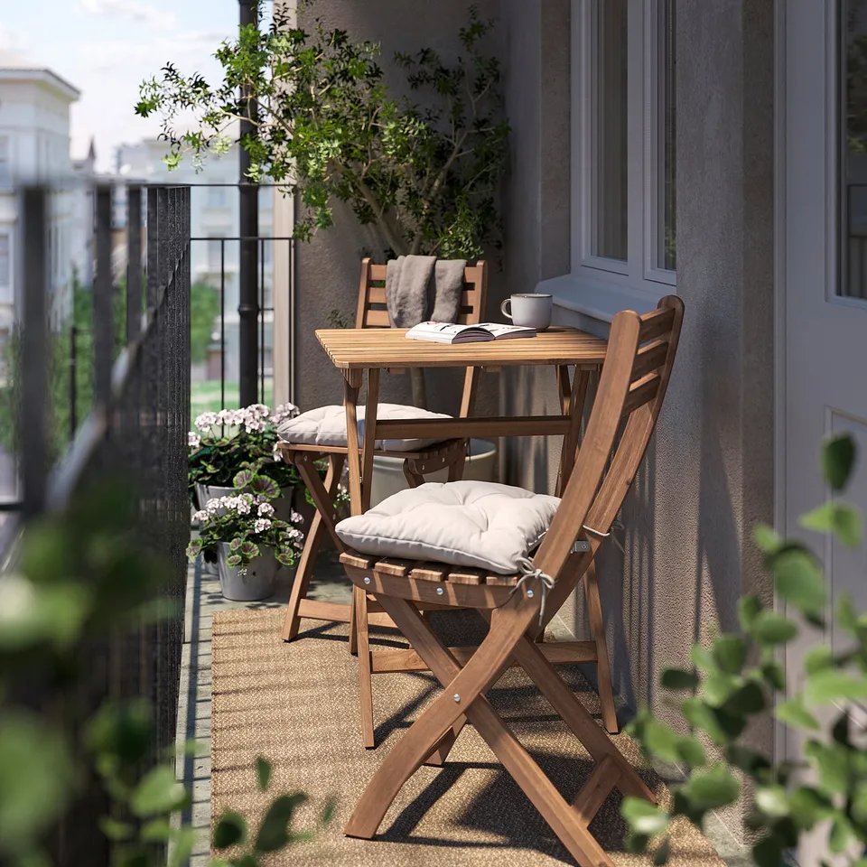 Cosy outdoor balcony with brown ASKHOLMEN wooden table and chairs, white cushions, plant, and mug.