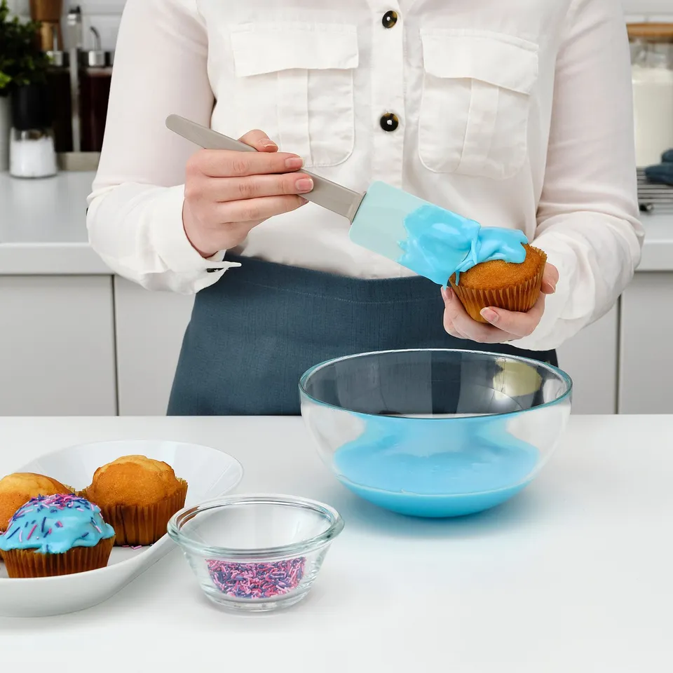 A person decorates cupcakes using a blue spatula with a small support to avoid countertop mess. Various baking items are present.