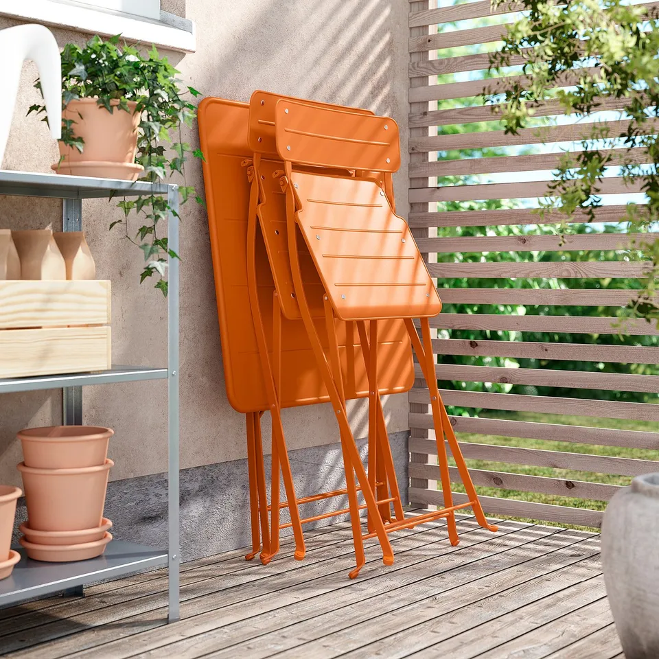 Three bright orange SUNDSÖ foldable chairs stacked neatly outdoors against a wooden fence.
