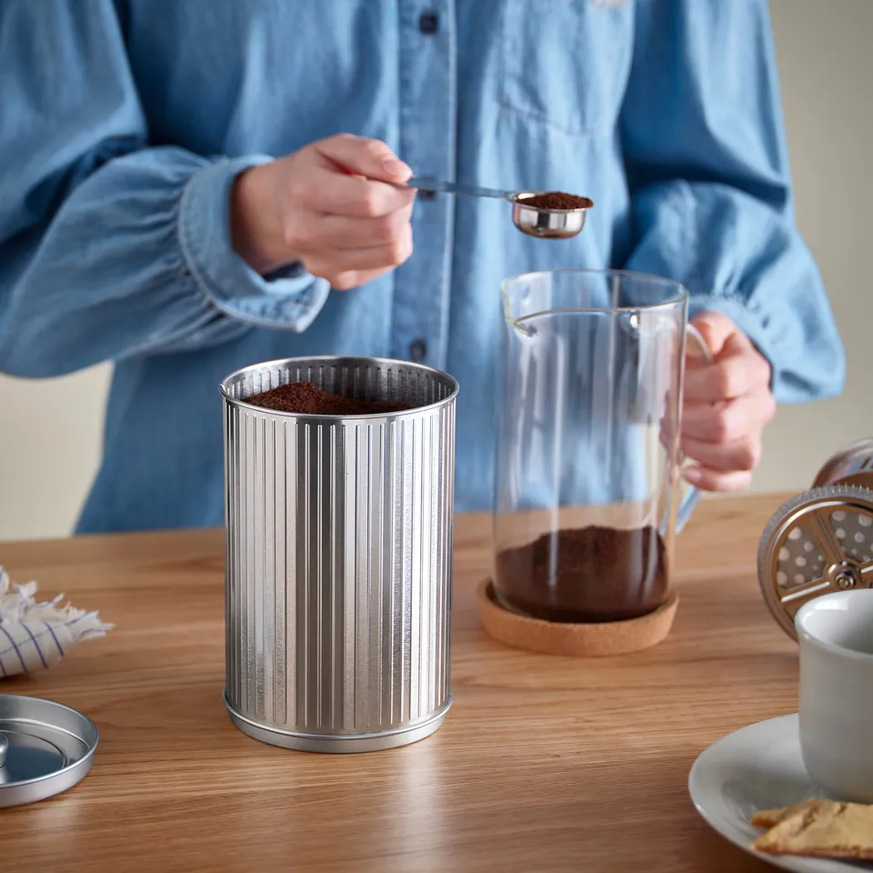 Person scooping coffee grounds into a metal container on a wooden table.