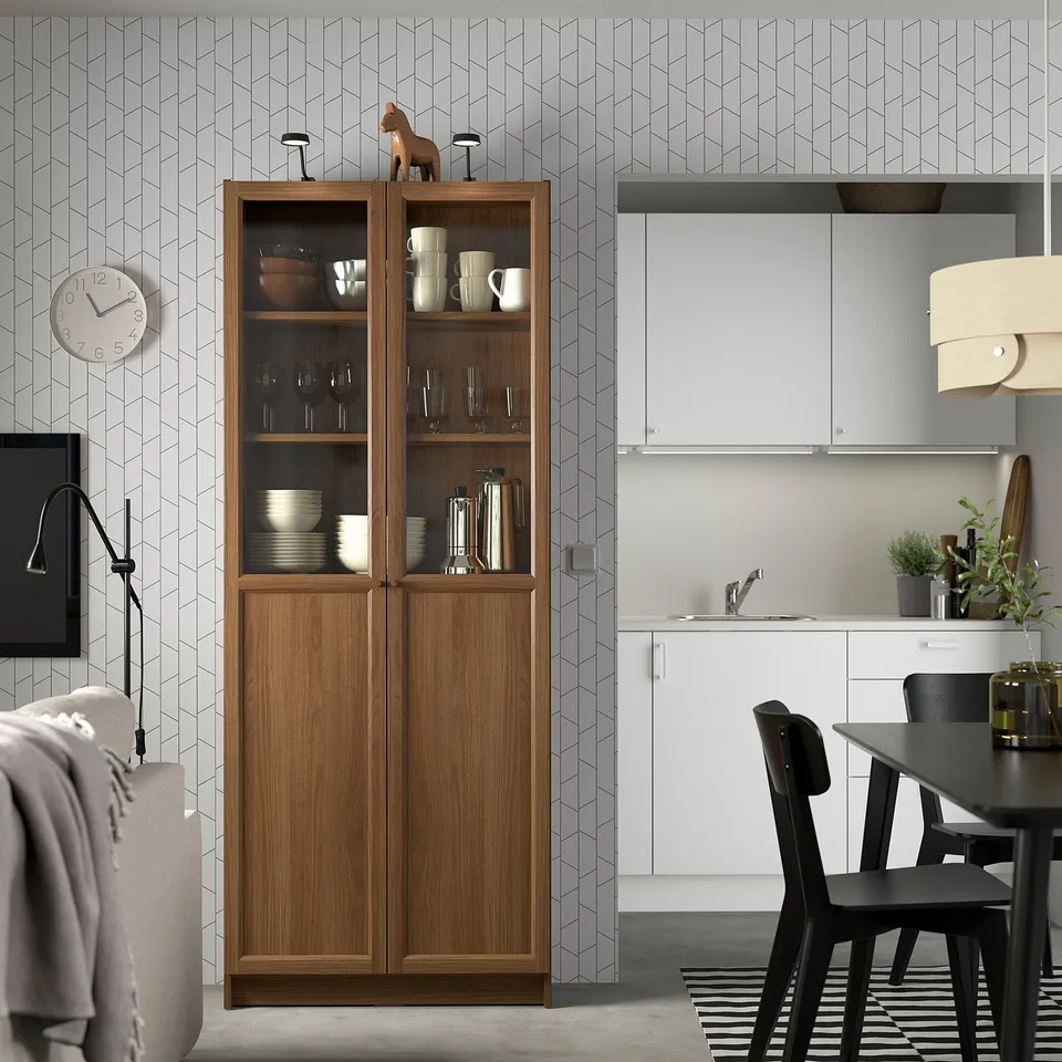 BILLY bookcase in natural finish glass and wooden doors displaying kitchenware. Its tall and narrow size fits perfectly in the modern kitchen setting.
