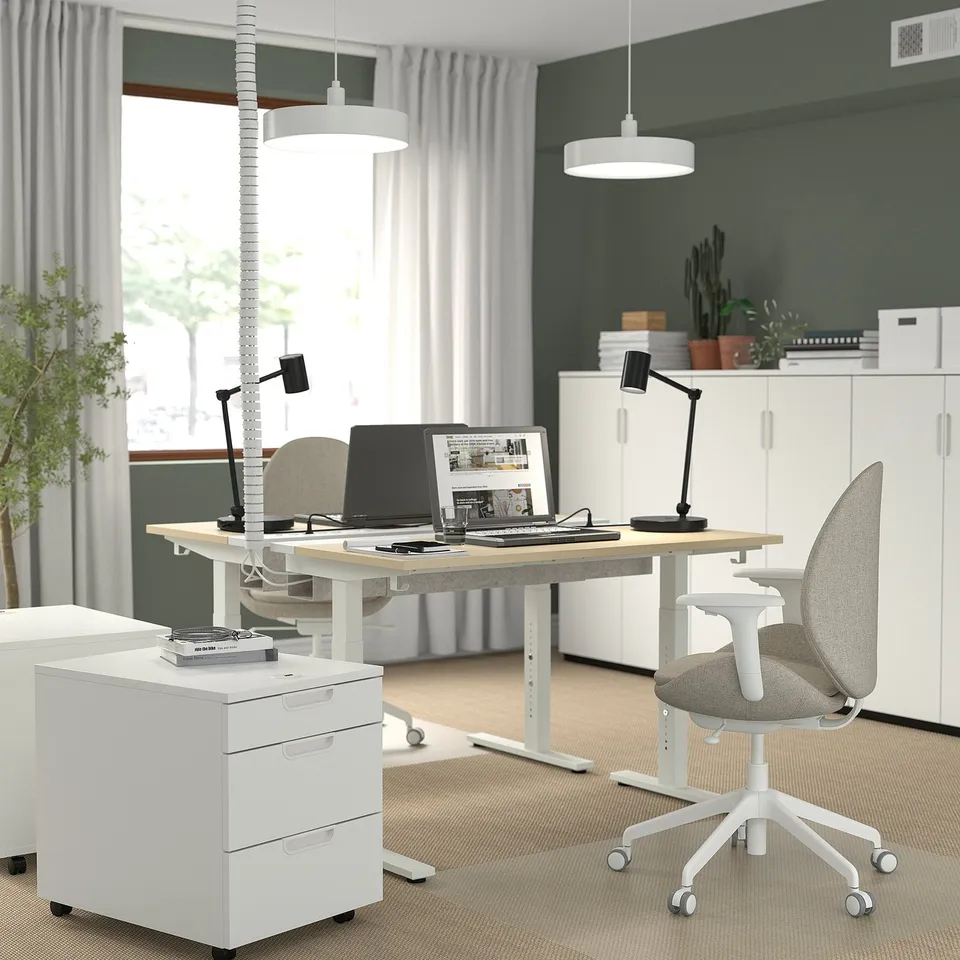 White height-adjustable desk with light wood top, metal base, lamp, filing cabinet, and grey chair.