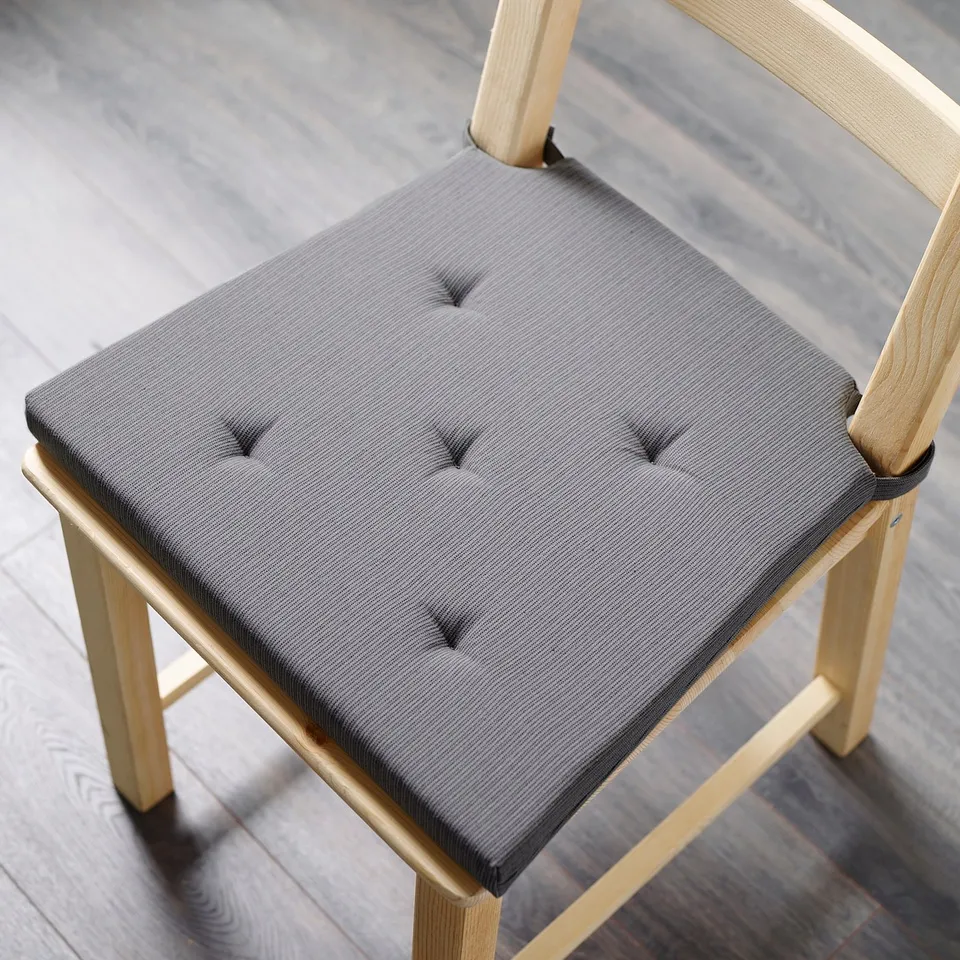 The JUSTINA chair cushion is grey, square, and fits perfectly on a light wooden chair. Its plush design adds comfort to dining settings.