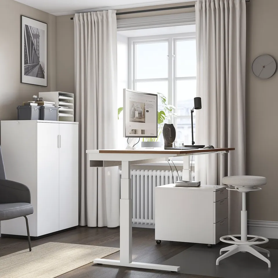 MITTZON white sit/stand desk. Modern, ergonomic design. Tall, with a wide top, larger than the nearby grey chair and white storage cabinet.