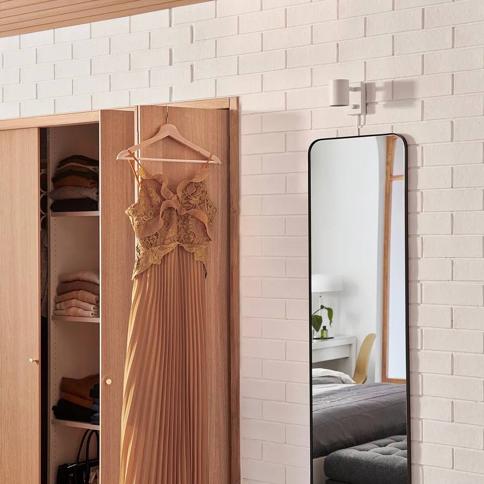 A beige dress hangs near a wooden closet with a mirror attached. The mirror has a black frame and is set against a white brick wall.