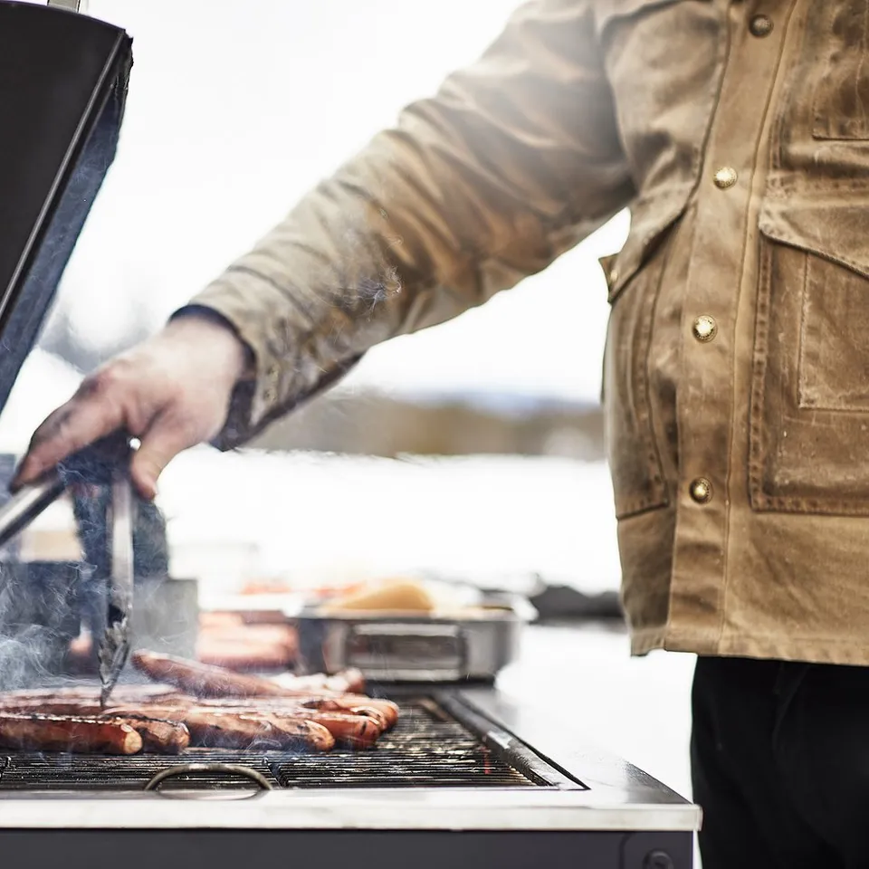 Person grilling sausages on a GRILLSKÄR charcoal bbq. Smoke rising, wearing brown jacket.
