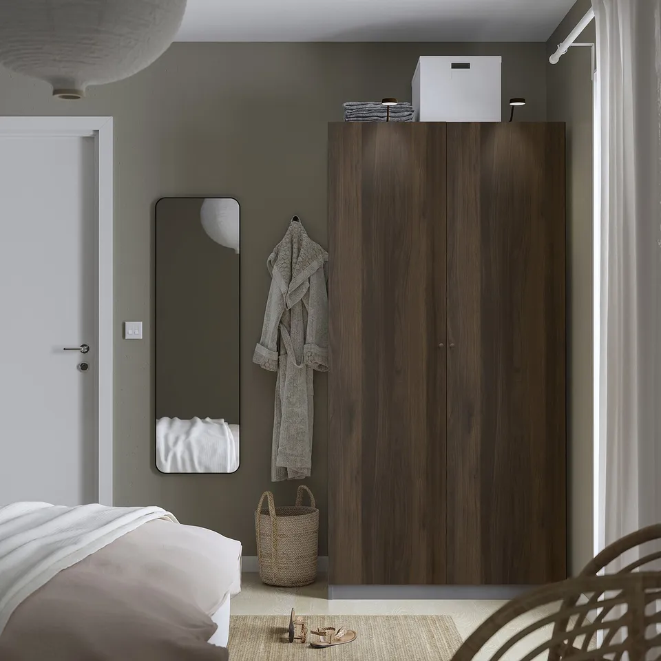 Modern bedroom tall brown PAX wardrobe, large mirror, white bed, wicker basket, and hanging robe. Wardrobe is larger than other items.