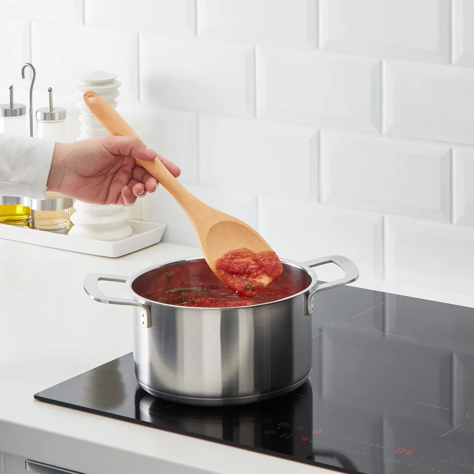 A wooden cooking spoon is stirring red sauce in a silver saucepan on a stovetop.