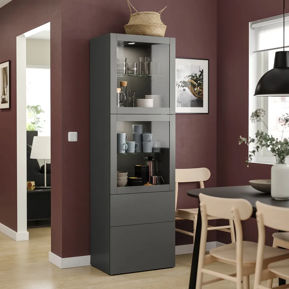 Display cabinet with glass doors storing cups and bowls. Fixed to wall with dark red walls, light wood chairs, and additional furniture.