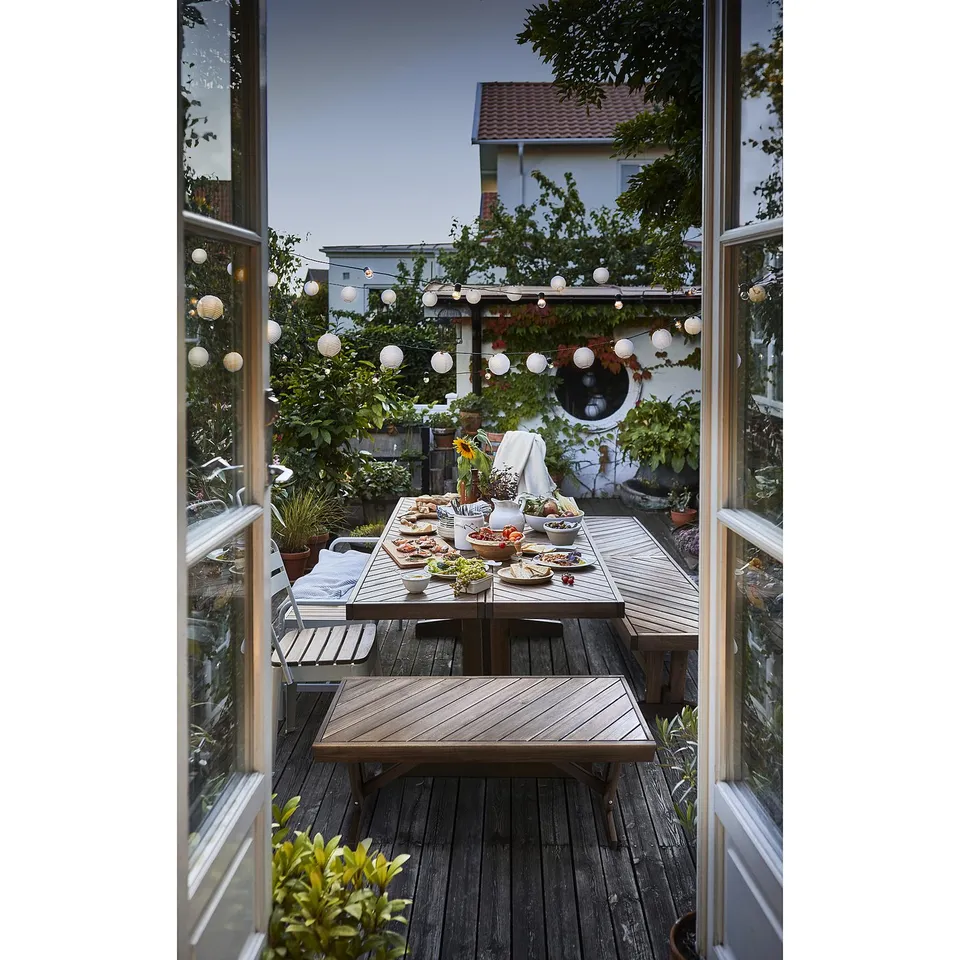 Outdoor wooden dining area with NORRMANSÖ bench. Round tables with food and hanging lights.