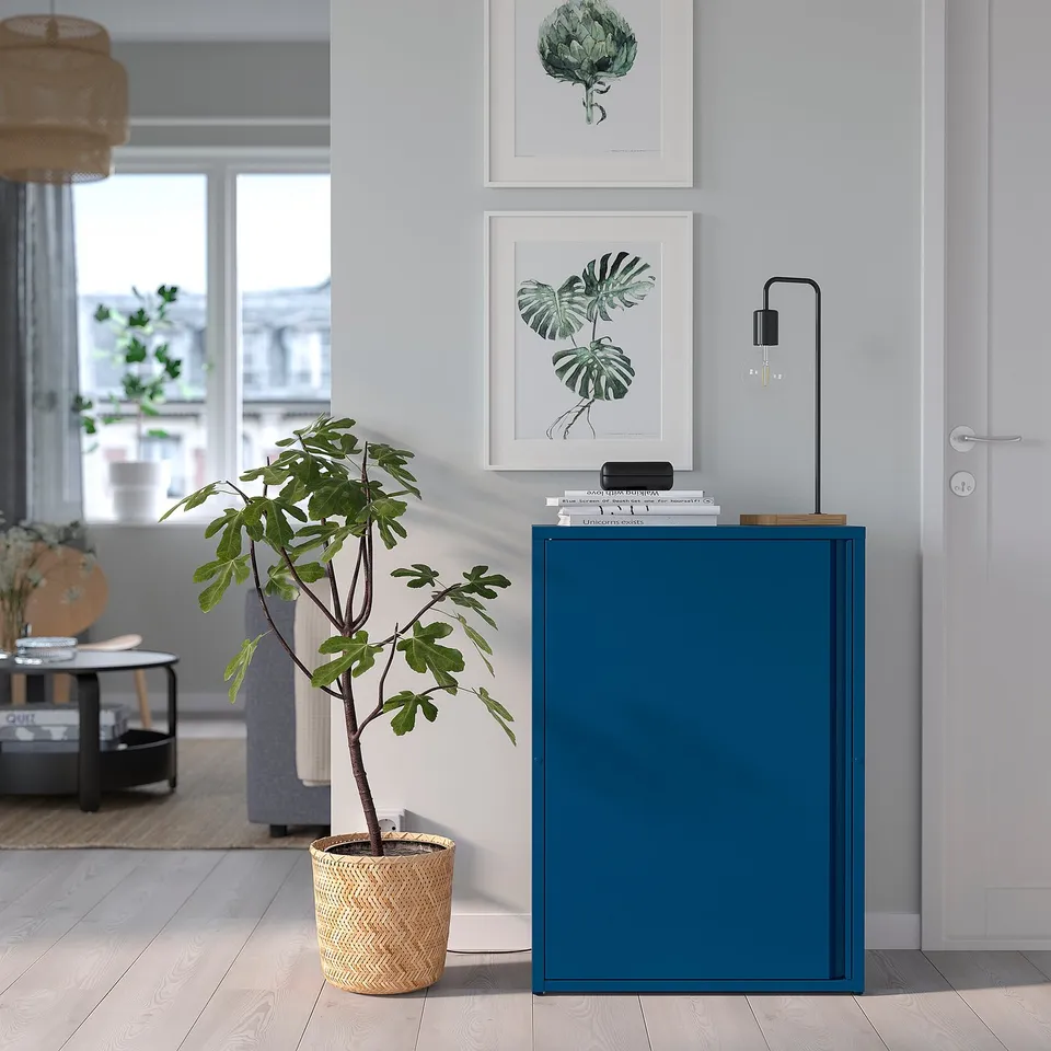 Modern room with teal SUNDSÖ cabinet, woven plant basket, wooden table, art prints, and large windows.