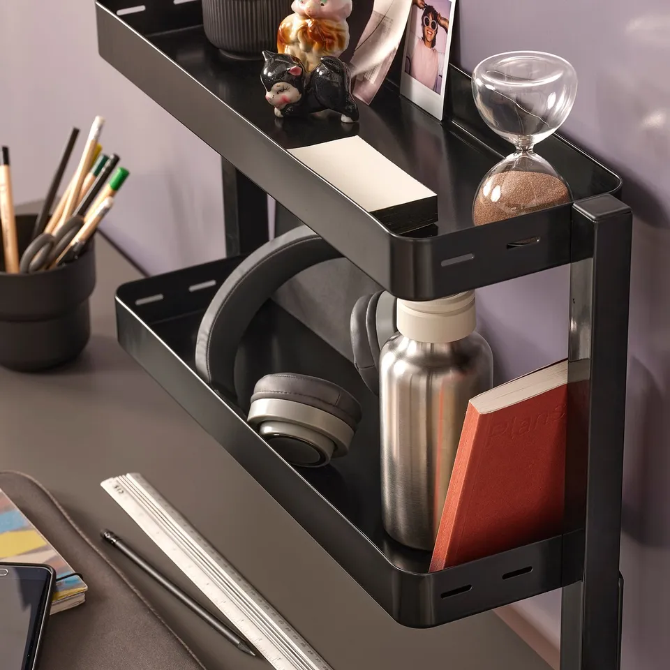 Black multi-level shelf organiser mounted on a table, holding various items including pens, a water bottle, headphones, and a notebook.