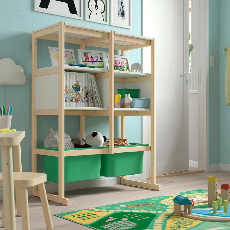A light wood-colored FLISAT storage unit with shelves holding books and toys. It stands near a door and has green plastic bins at the bottom.
