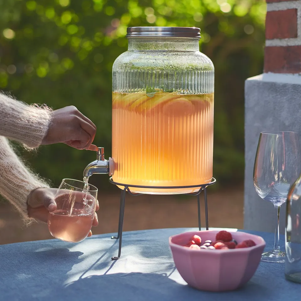 A person pours orange drink from a clear glass jar with a tap on a black stand, next to an empty wine glass and a bowl of berries.