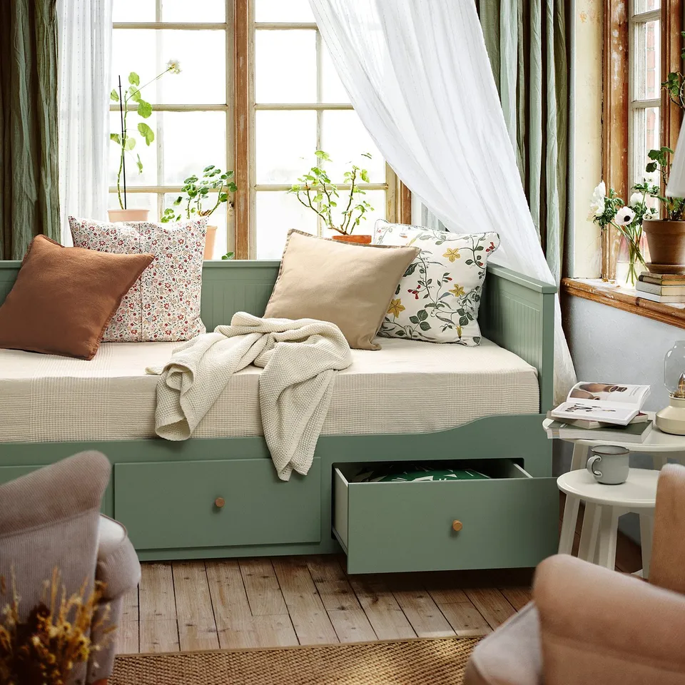 A grey-green day-bed with large drawers, set by a window. Its used as a single bed, with drawn curtains and a cosy blanket on top.