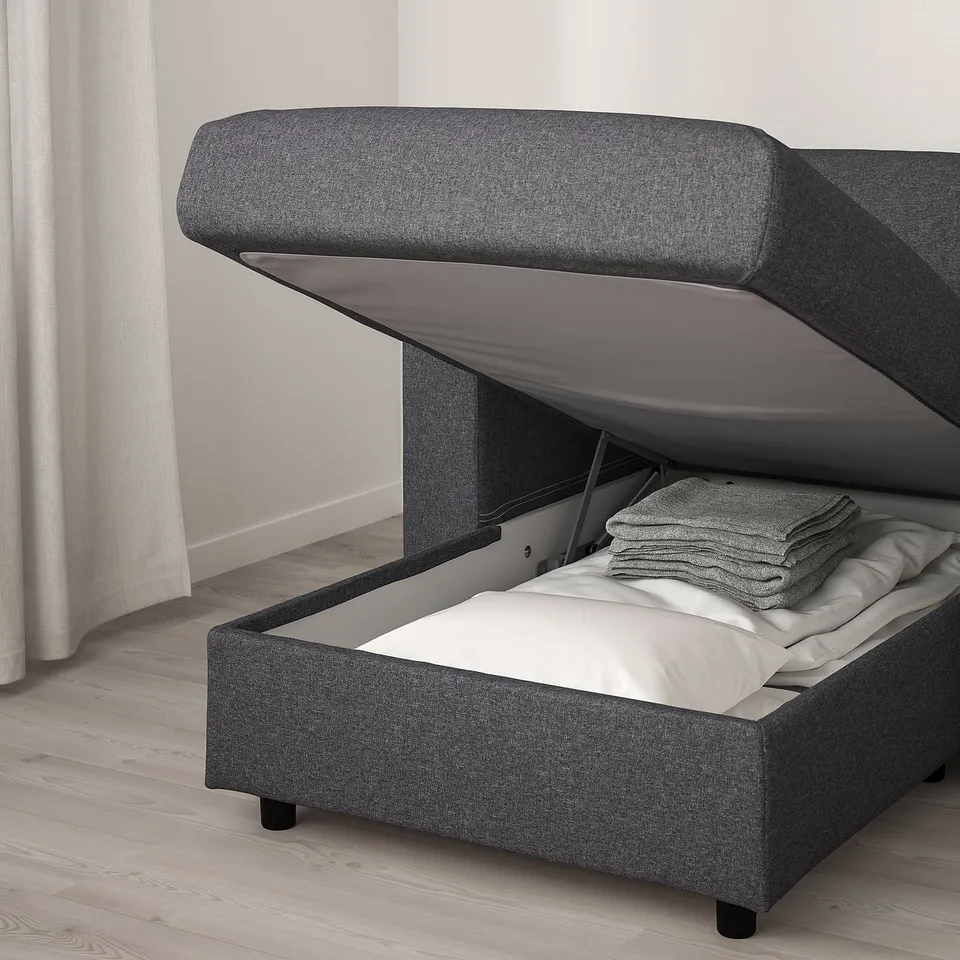 A grey upholstered sofa-bed with storage. The lid is open, showing stacked items inside. The bed is partially visible with white linens.