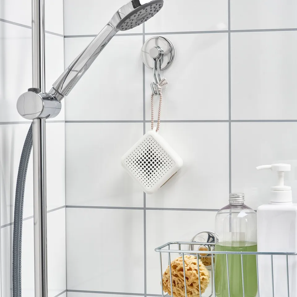A waterproof white speaker hangs in a bathroom by a hook, attached to a shower head.