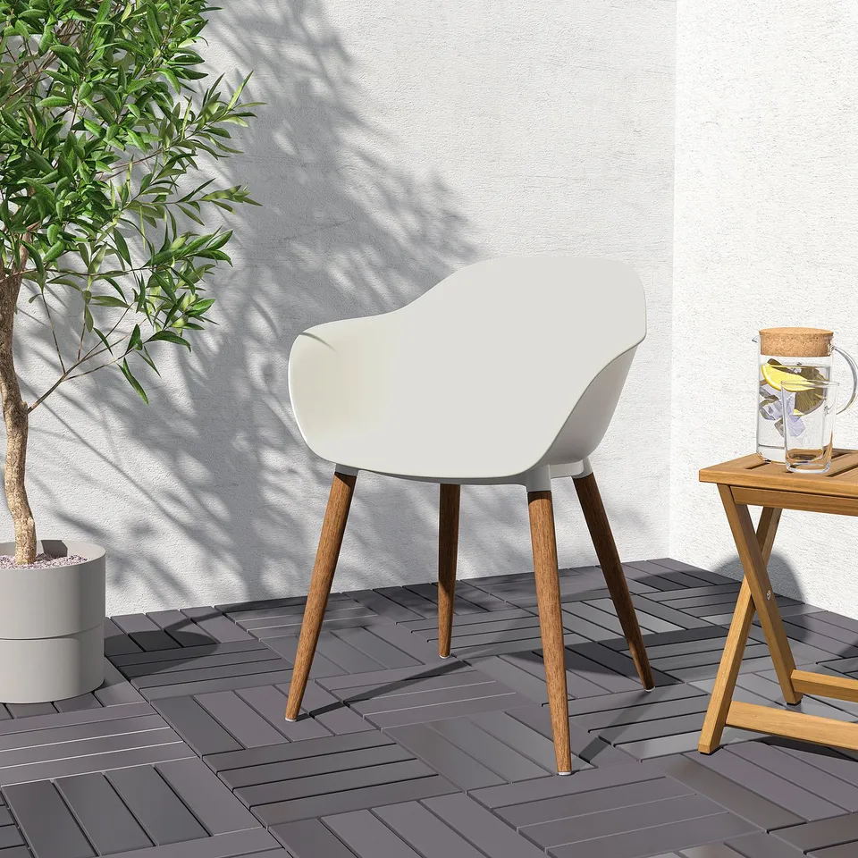 Modern outdoor scene: white GRÖNSTA chair with wooden legs by a small table holding a drink, near a large plant against a light wall.