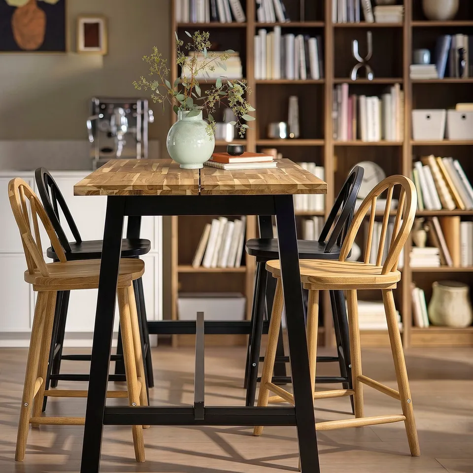SKOGSTA bar table with acacia wood top, black metal legs. Matching chairs with wooden seats in cosy setting.