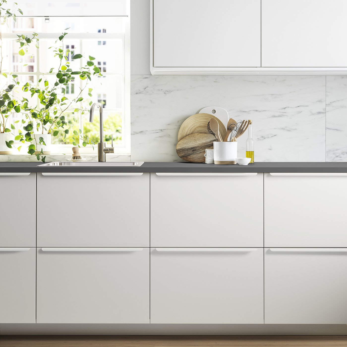 Modern white JÄRSTORP kitchen with marble backsplash, sleek cabinets, dark counter, utensils, plants, sink, and window.