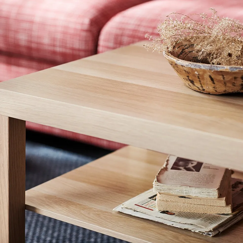 Light wooden LACK coffee table with stacked books and dried plants bowl next to red fabric sofa.