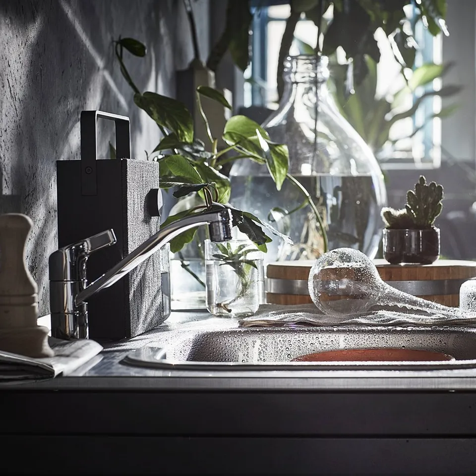 Modern kitchen sink with stainless steel tap, glass jars, and potted plants. Sunlight streams through window.