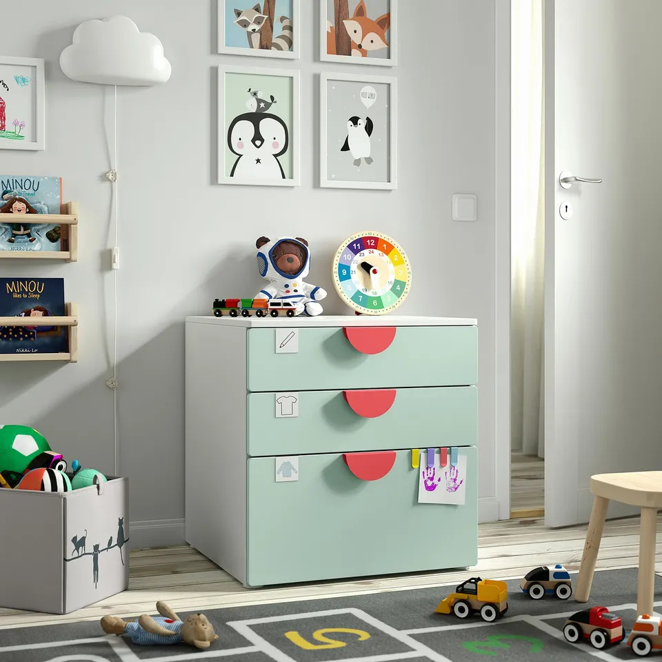 Kids room with small green chest, red-handled drawers, a colourful clock, and toy-filled bookshelf, surrounded by playmat and cars.