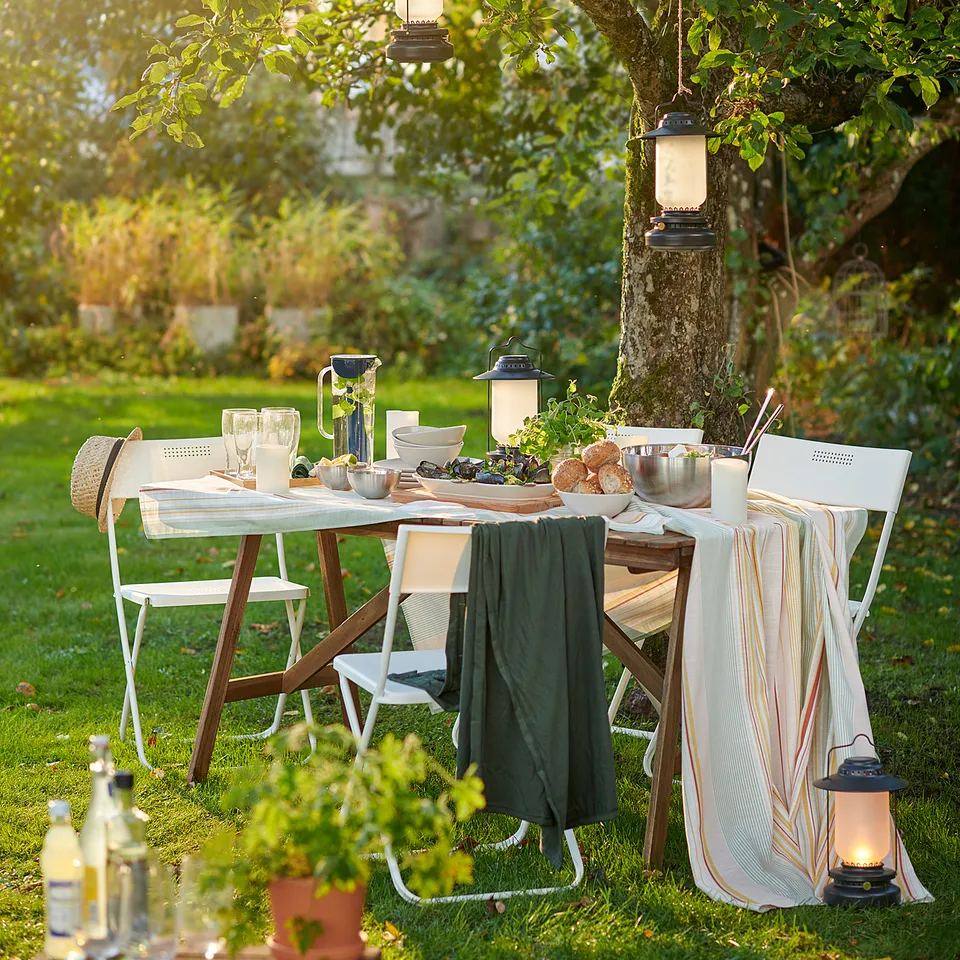 Outdoor table under a tree with lanterns. Set with green cloth, plates, glasses, and food. Two white chairs and a drinks table nearby.