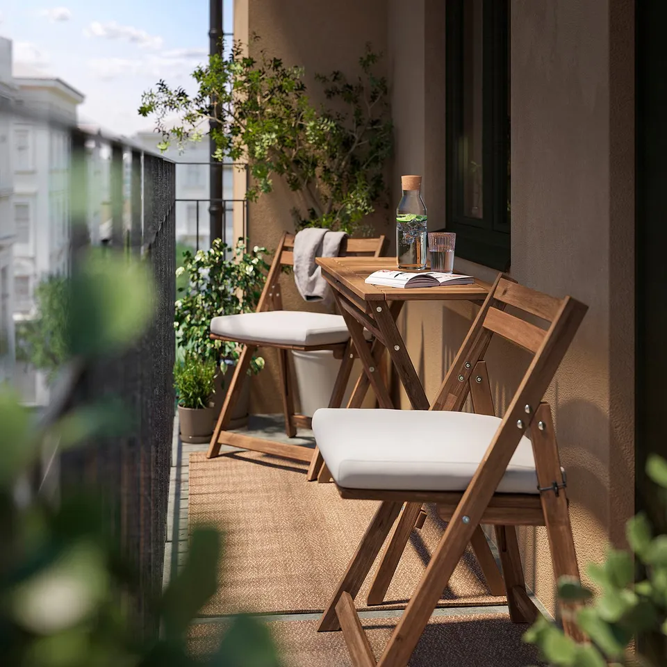 Small balcony setup with foldable wood table and chairs, white cushions. Urban view, greenery, and a relaxed vibe.