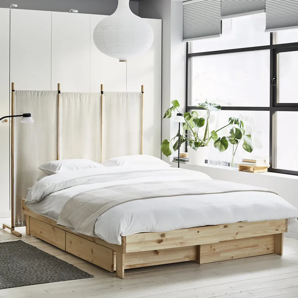 Minimalist bedroom with a light wooden GLAMBERGET extendable bed, white linen, and a large window with plants.