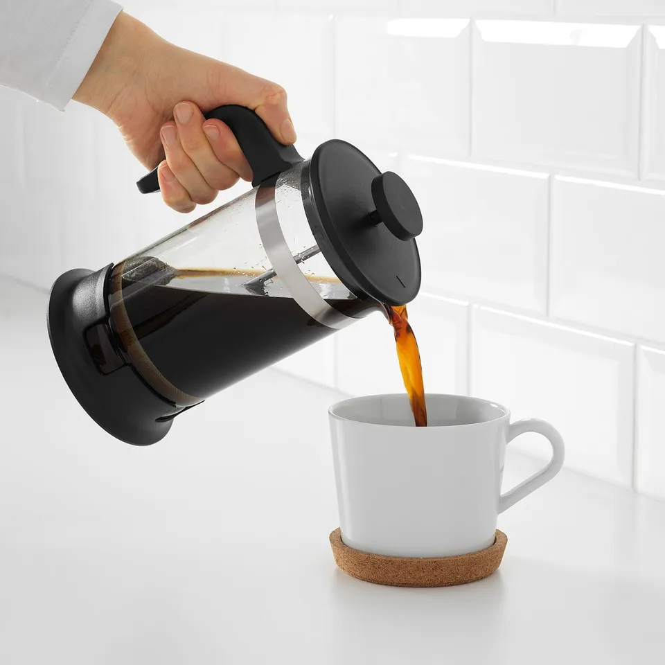 Person pours freshly brewed coffee from a cafetière into a white mug on a cork coaster.