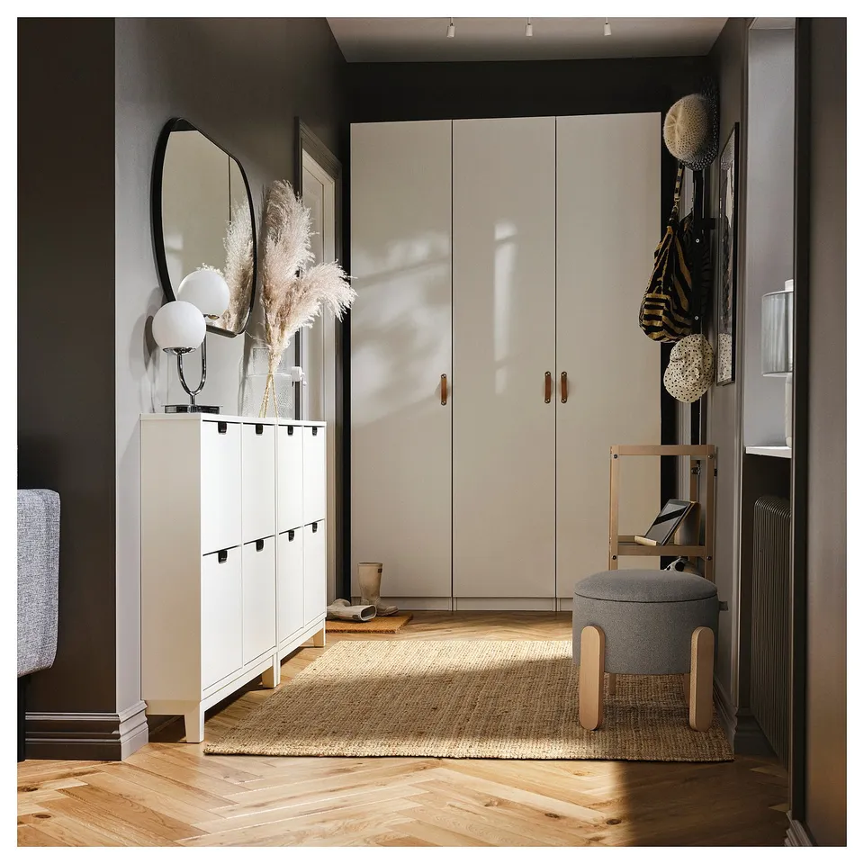 Modern entryway with white cabinets, mirror, dried flowers, and grey storage ottoman on a wooden floor.