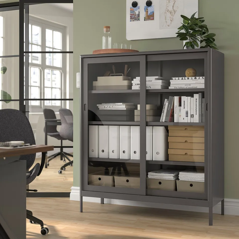 IDÅSEN black storage cabinet with glass doors in office, holding books and decor, saves space.