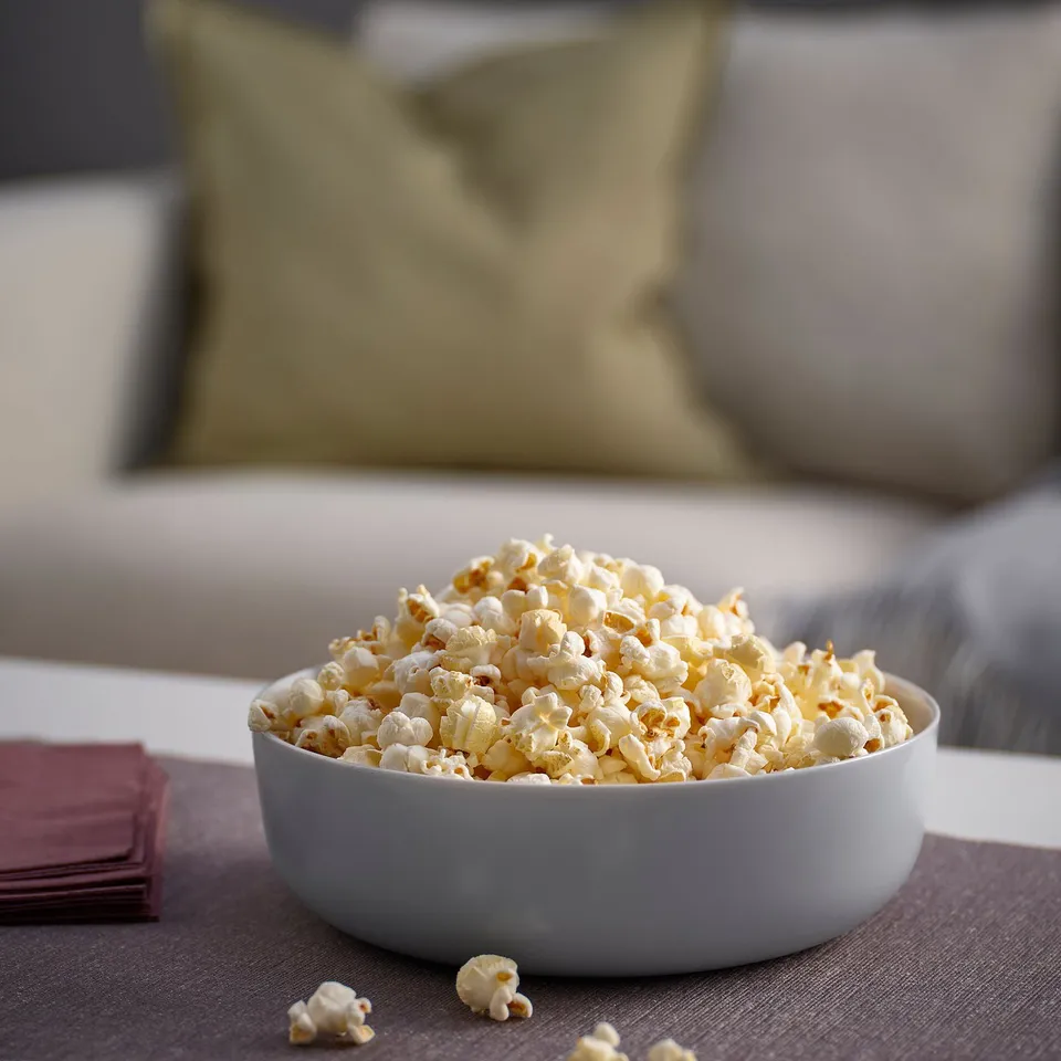 A white tempered glass bowl holds popcorn, placed on a sofa with cushions and a partially visible person.