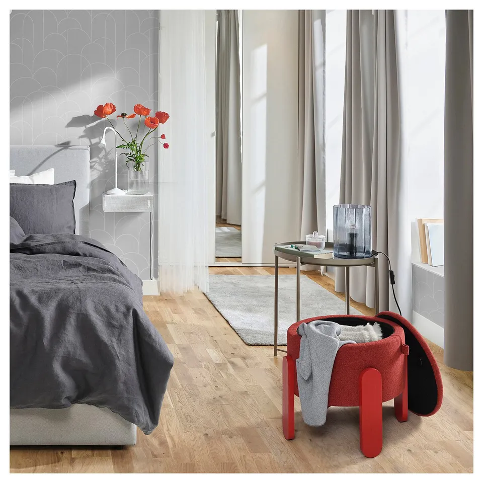 Modern bedroom with FÖRLUNDA red ottoman offering seating and storage, next to a bed with grey bedding and a bedside table holding red flowers.