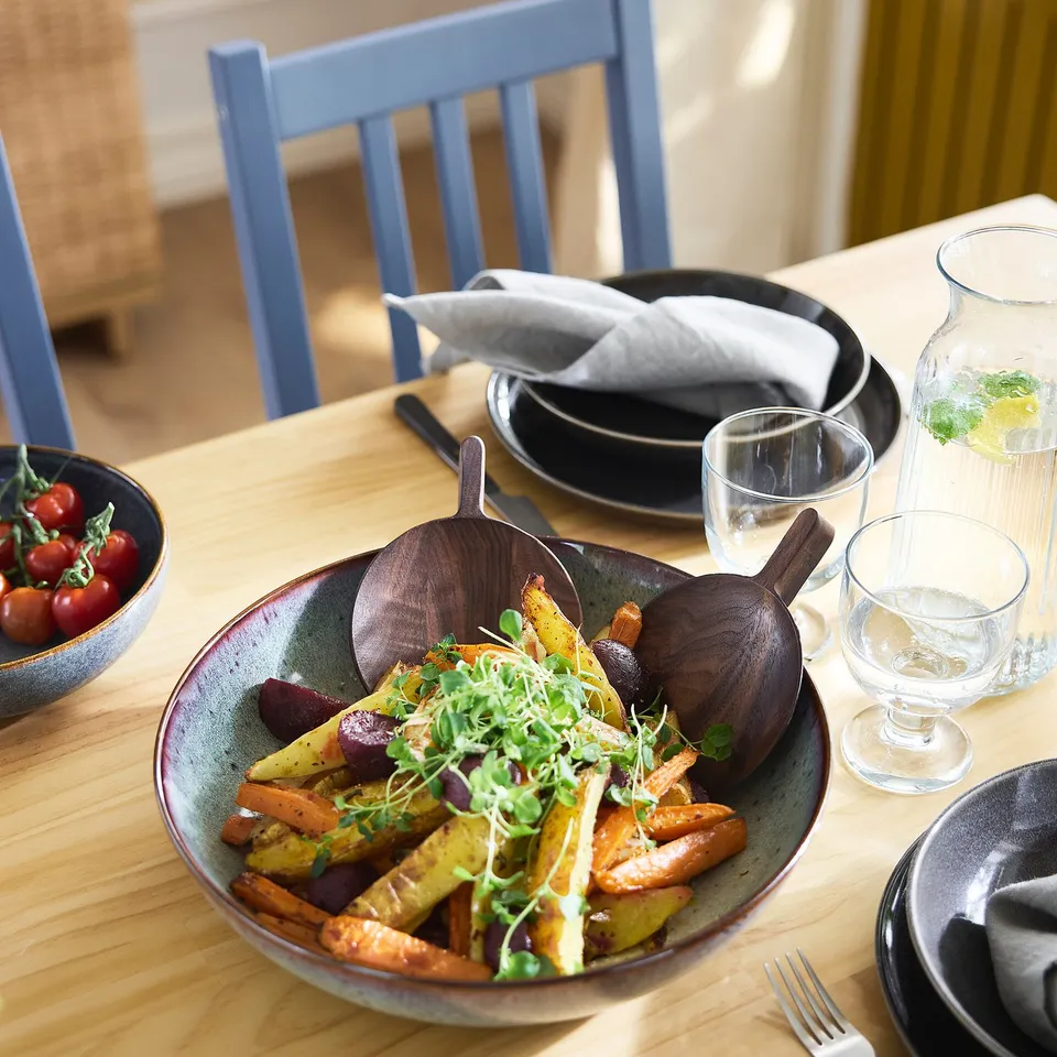 Wooden dining set with roasted veggies, blue chairs, glasses.