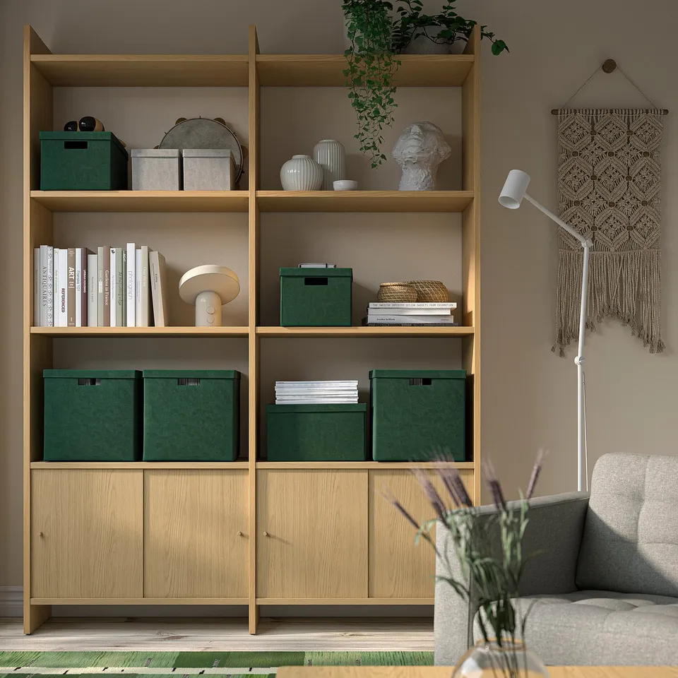 Wooden bookshelf with green storage boxes, books, vases, and macrame decor. Grey sofa and lamp nearby.