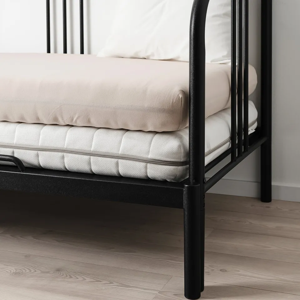 Black metal bed with beige mattress and pillows on wooden floor.