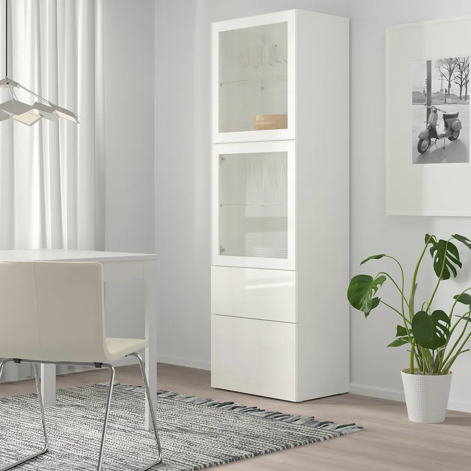 White BESTÅ cabinet, glass doors, chair, rug, plant. Minimalist room decor.