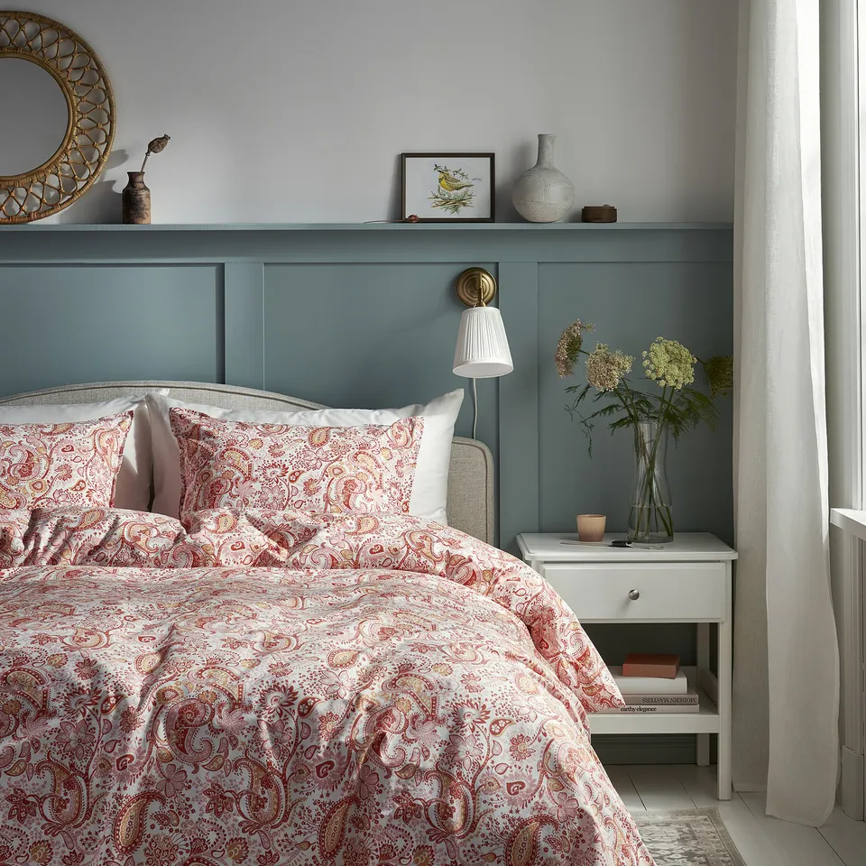 Bedroom with paisley bedspread, teal headboard, side table with flowers and candle, shelf with mirror and lamp, grey walls, curtains.