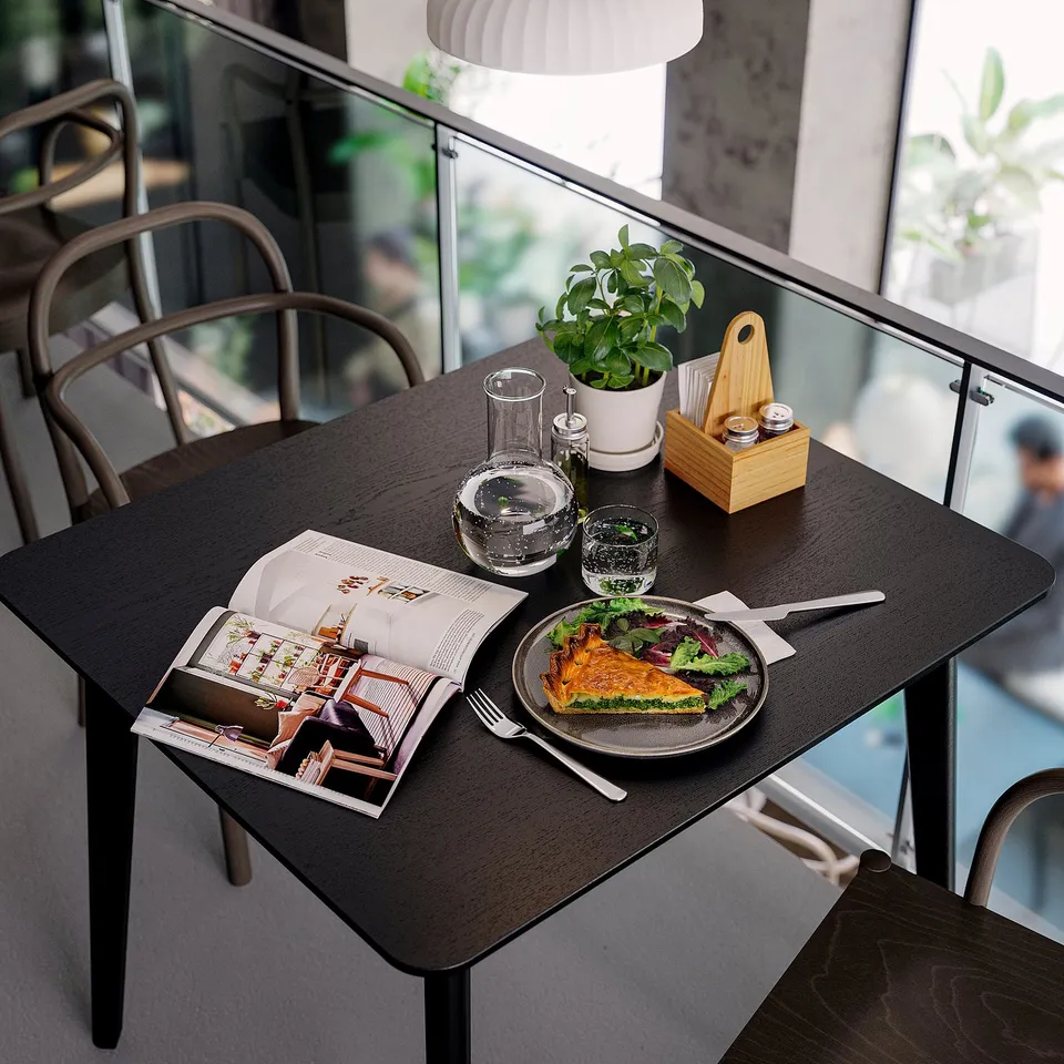 Small dark wooden dining set with plate of quiche, open magazine, glasses, and plant by window.
