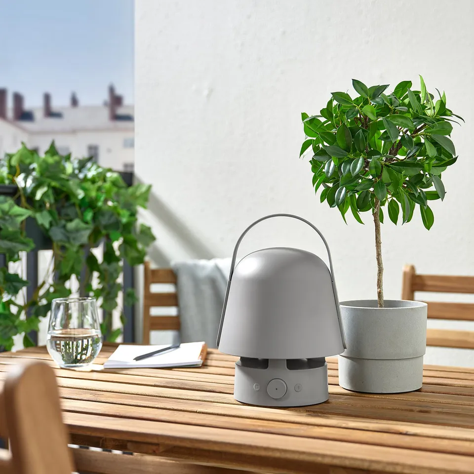 Outdoor table with grey VAPPEBY speaker lamp, plant, glass, and notepad. Lamp is small, round, with a handle on top.
