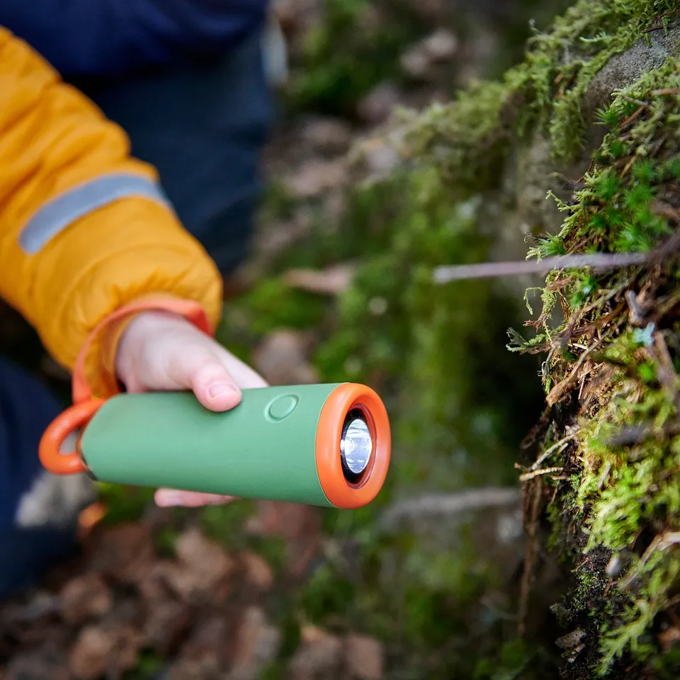 Person holding green and orange led torch with string in mossy outdoor area. The torch is hand-driven, lights up when string is pulled.