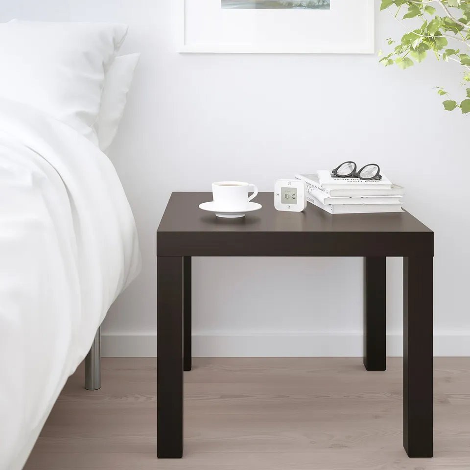 Small LACK side table in bLACK beside a bed. Holds a cup, alarm clock, books, and glasses. Simple, square design with four thin legs.